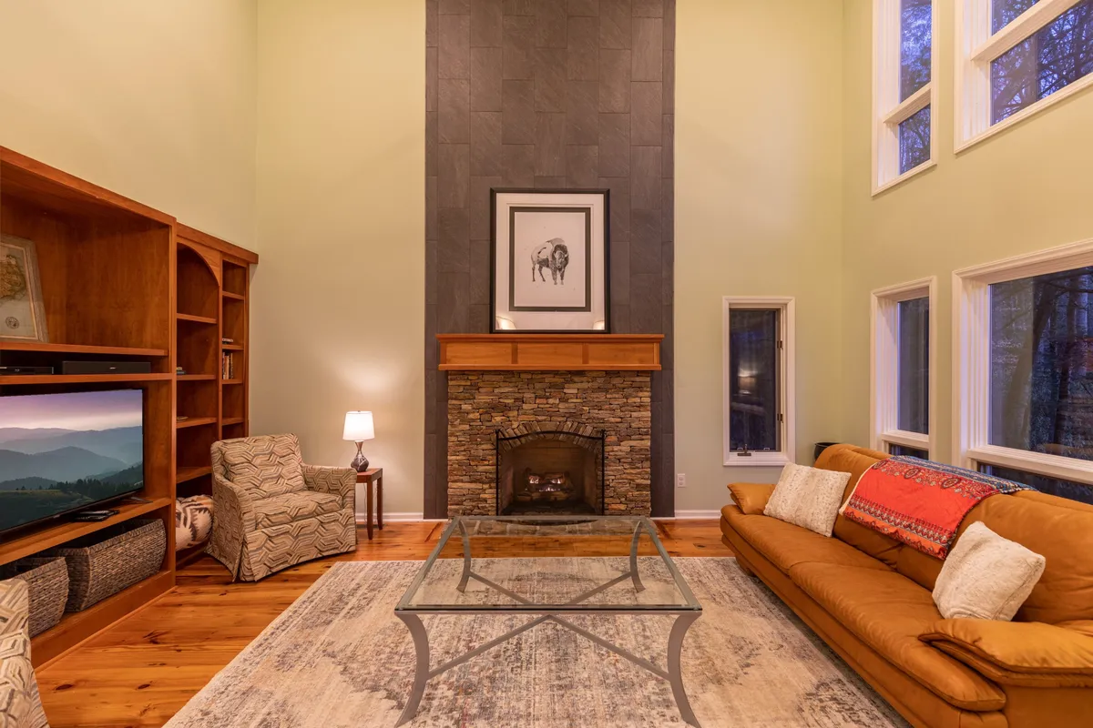 A spacious living room with a stone fireplace, large windows revealing lush forest views, a comfortable sofa, and tastefully arranged artwork. The room is bathed in natural light and exudes warmth and welcome.