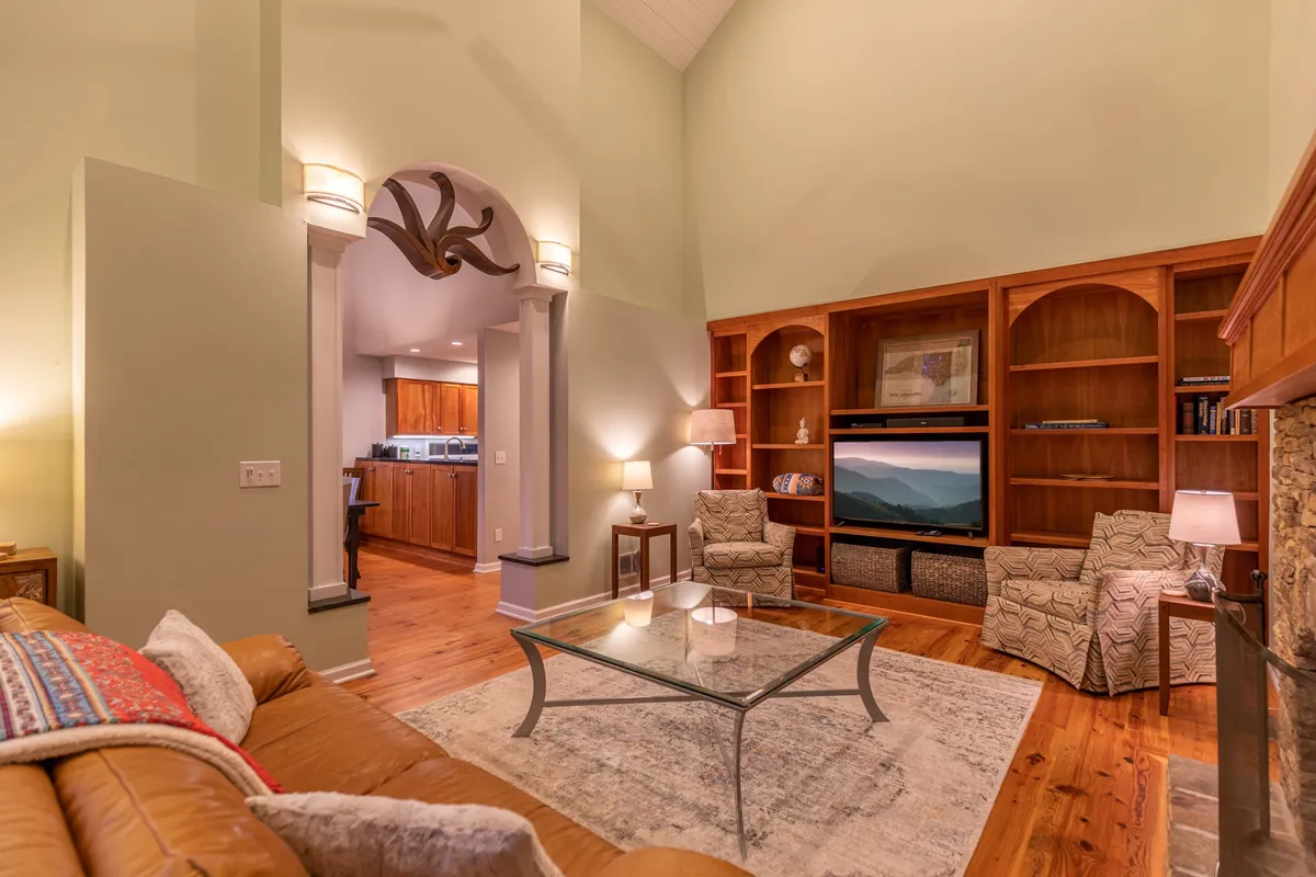 An intimate living area featuring a leather sofa, glass coffee table, bookshelf, TV, and armchairs. The arrangement encourages relaxation, socializing, and movie nights in a comfortable, welcoming environment.