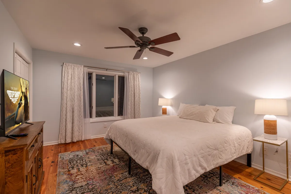 A cozy bedroom with a large, inviting bed, a window bringing in natural light, a nightstand with a lamp, and a television for evening relaxation. The soft color palette sets a restful mood.