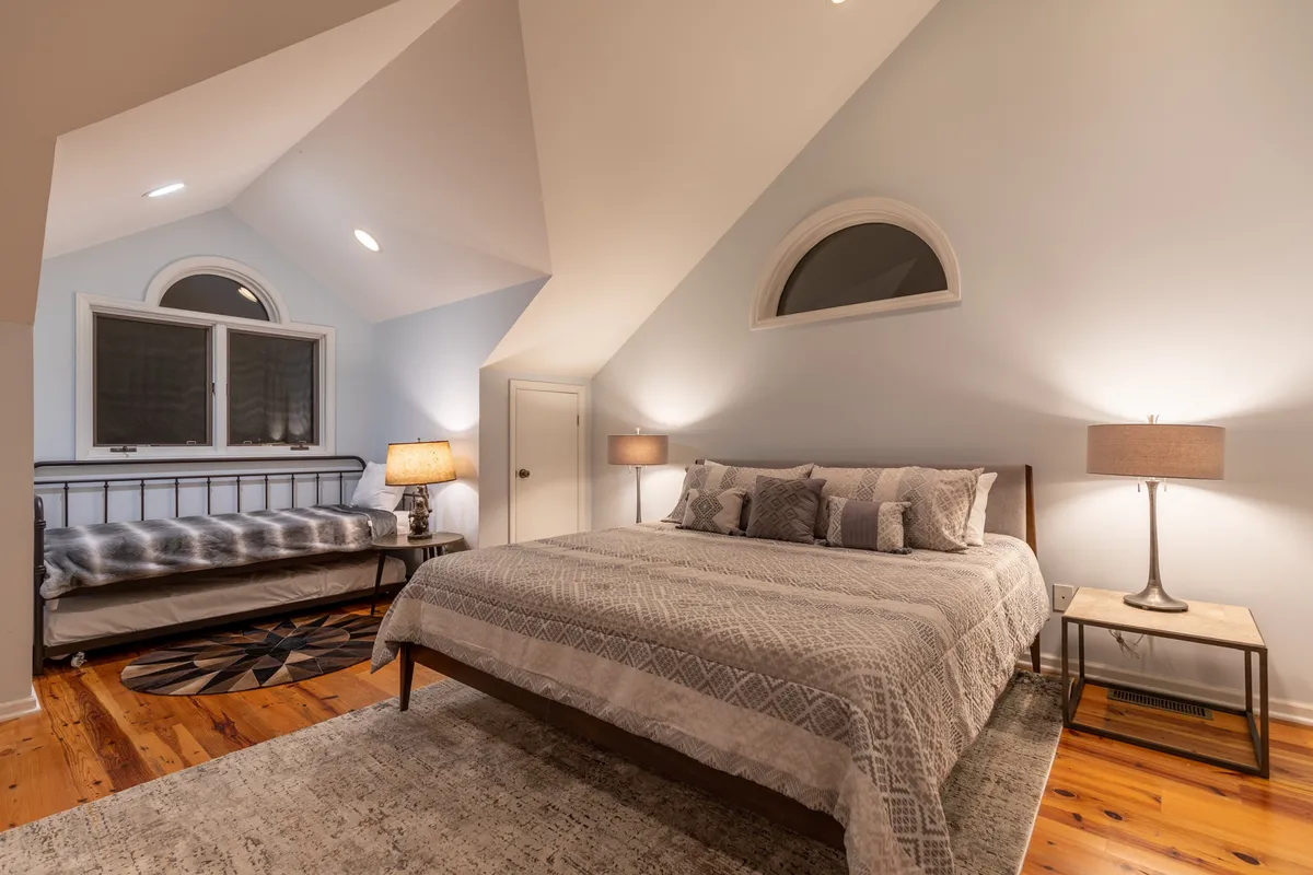 A spacious and inviting bedroom featuring a king-sized bed with decorative pillows, bedside lamps, a window with soft drapes, and a daybed against the wall. The room feels airy and bright, ideal for relaxation or quiet moments.