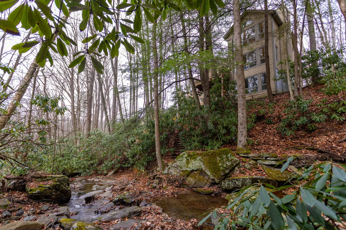 A tranquil exterior shot of the house nestled among trees, with a flowing creek and natural rock formations visible. The environment is peaceful, emphasizing the proximity to nature and the creek.