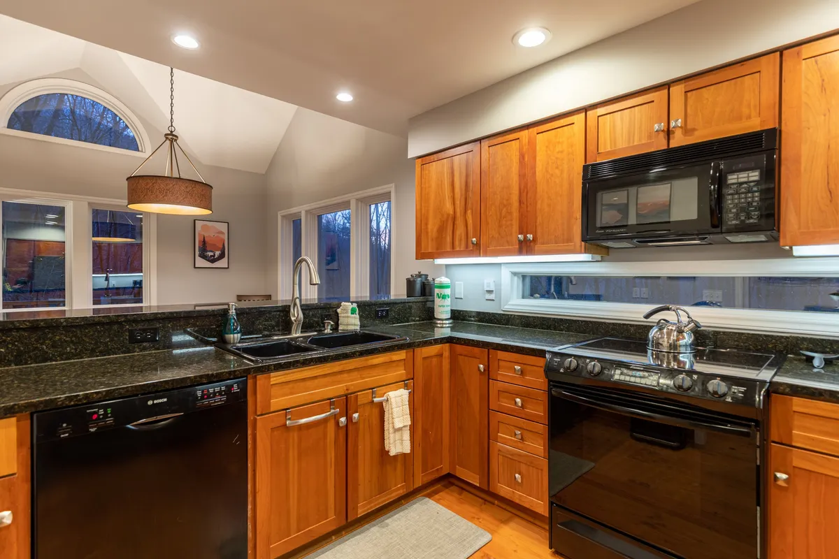 This image features a spacious kitchen highlighted by a large island with black granite countertops, modern appliances, wooden cabinetry, and generous windows that flood the space with natural light. The layout is clearly designed for group cooking and entertaining, with ample prep space and a cozy, inviting atmosphere.