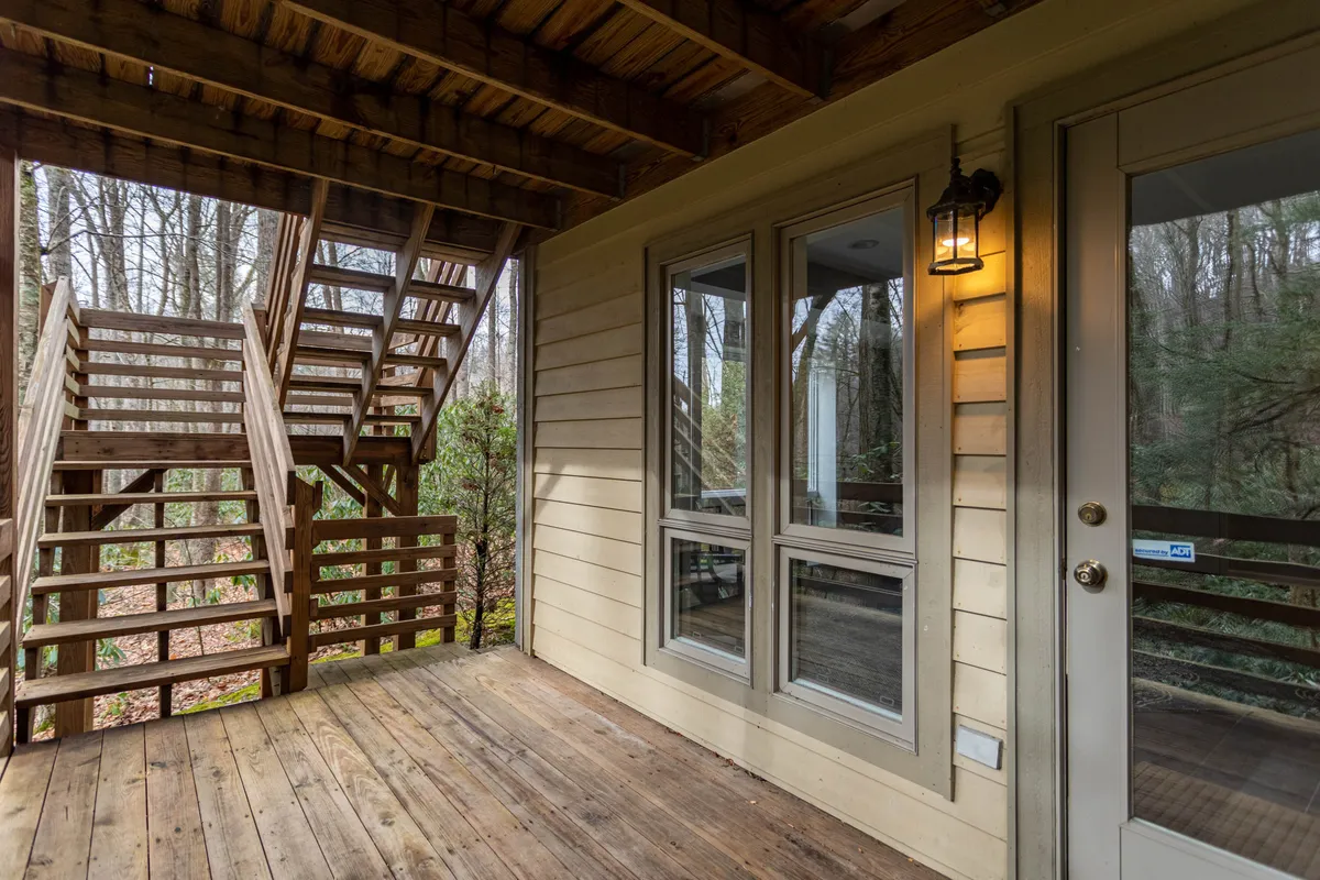 This image captures a welcoming interior view with prominent wooden stairs, a front door, and expansive windows that frame nature views. There is a clear connection to the outdoors, with sunlight streaming in, and a porch area suggested by the layout. The ambiance invites guests to imagine starting their day in the mountains and returning to comfort.